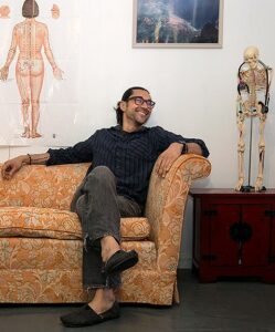 A picture of Frank Coughlin, MD. He is a white man with long dark hair pulled back, wearing glasses. He is looking to the left and is smiling. He is sitting on a gold, flowered couch with his legs crossed. Behind him on the wall is an anatomy poster of the back of a woman, and a teaching skeleton.