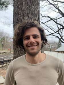 A picture of Jesse Marshall, a white man with short curly hair. He is standing in front of a tree trunk, he is smiling, and he is wearing a tan shirt.
