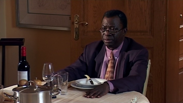 Still from Balfua Bakupu-Kanyinda’s film Juju Factory. A Congolese man sits at a table, set with a bottle of wine, dishes, and cutlery. He is wearing a suit with a purple button-down shirt.