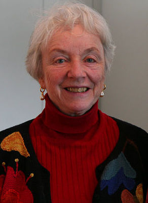 Color headshot of Madeline M. Kunin. Her grey hair is cropped short. She is wearing a red turtle neck and a multi-colored cardigan.