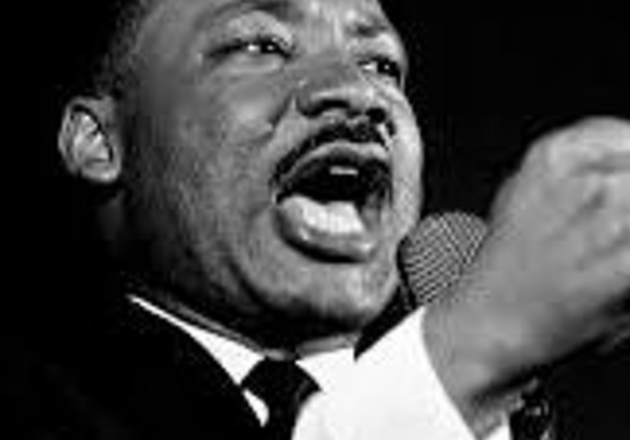 A black and white headshot of Martin Luther King Jr, speaking into a microphone and gesturing with his other hand.