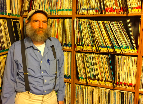 A photograph of Norm Stockwell who is a bearded man standing next to many shelves filled with vinyl records. He is wearing a baseball hat, a blue button down shirt, and suspenders.