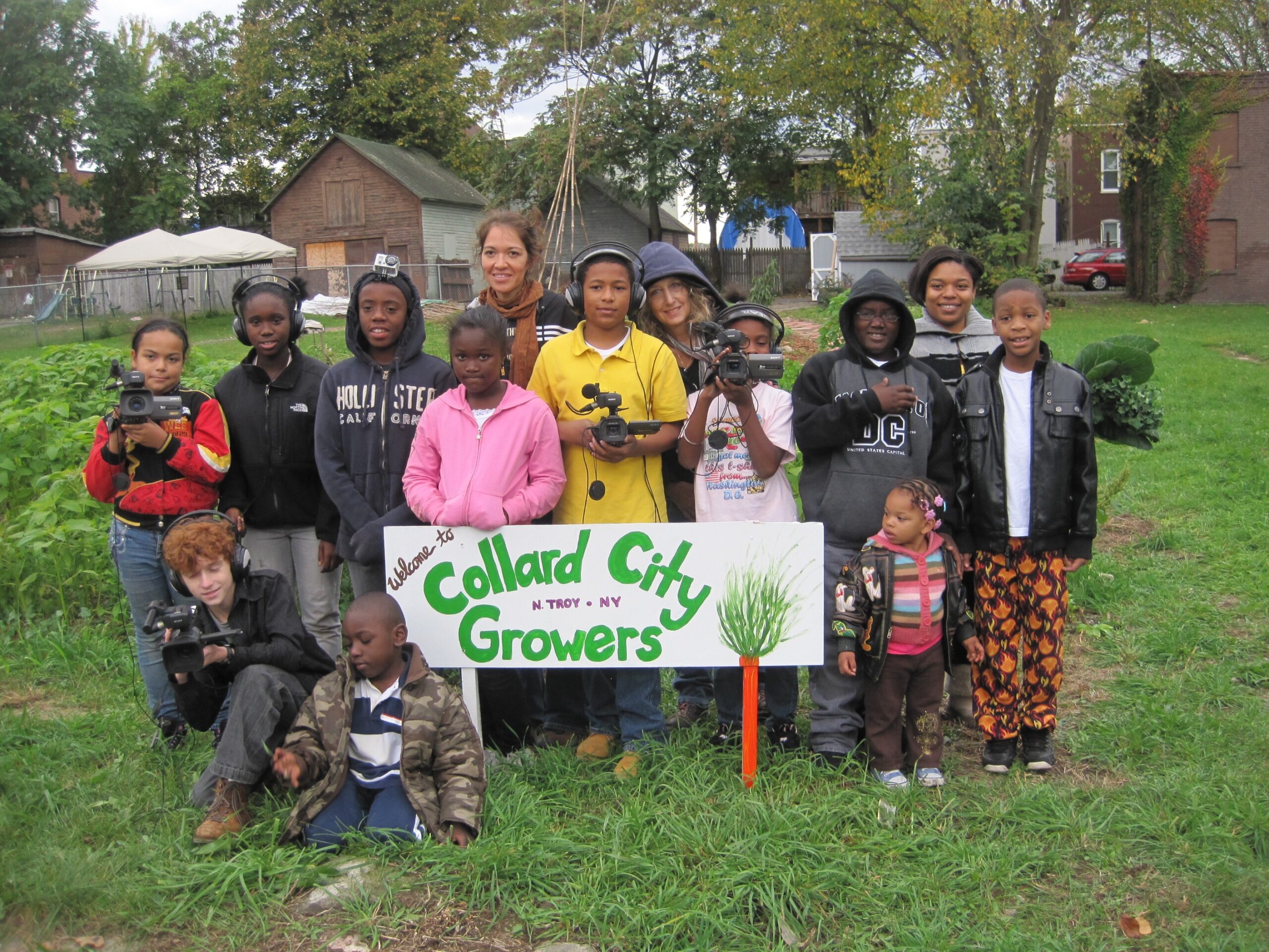 A group of youths, some of them wearing headphones or holding cameras, pose behind a sign that reads "Welcome to Collard City Growers, N. Troy, NY."