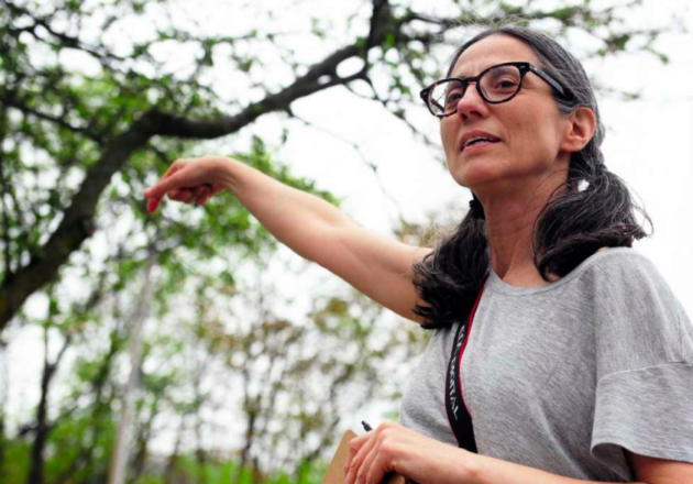 A picture of Maria Michails pointing to a tree in the background. She has her hair in pigtails, she is wearing a gray t-shirt and glasses.