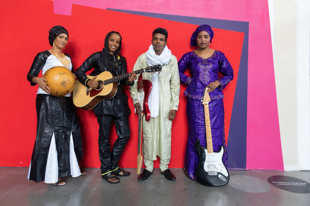A photo of Les Filles de Illighadad holding instruments (2 electric guitars, acoustic guitar, calabash) in front of a bright wall with red, dark purple, and pink. They are 4 Nigerien people: 3 women with hair in wraps, 2 of them in dresses, 1 man in a tunic. 