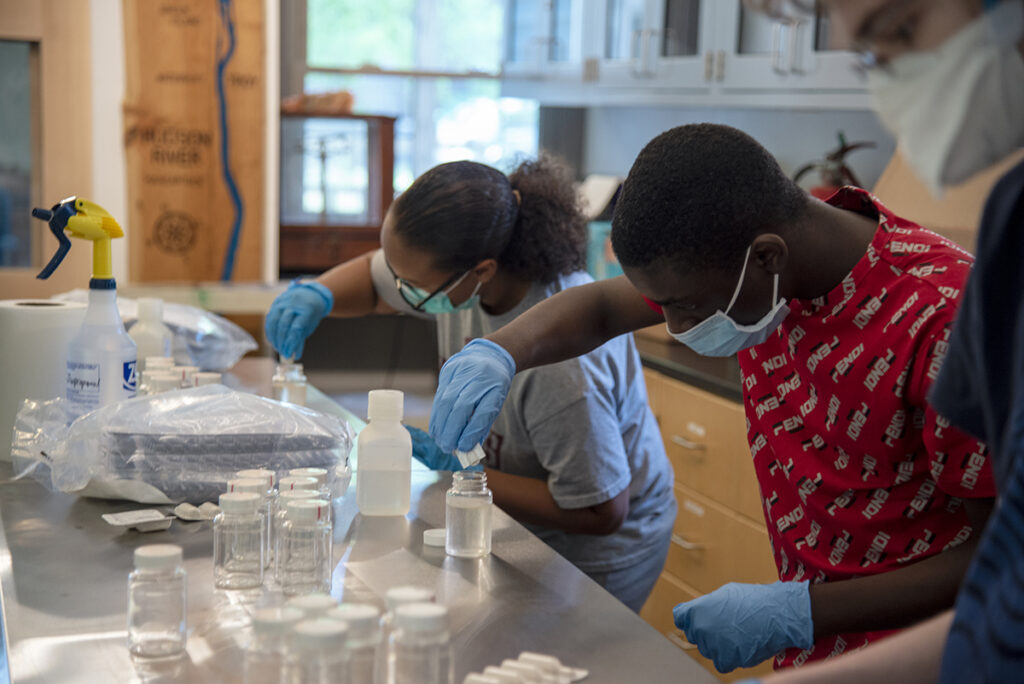 Three young people, all wearing surgical masks and gloves, work with vials of clear liquid in the NATURE Lab. 