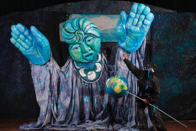 Large blue statue with an actor holding a globe on a stick and gesturing to it. The statue has two large hands lifted in the air and a face carved into it.