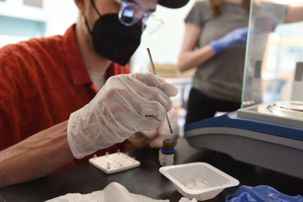 A light-skinned person wearing glasses, a black KN95 mask, and white vinyl gloves works with small tools in the NATURE Lab. 