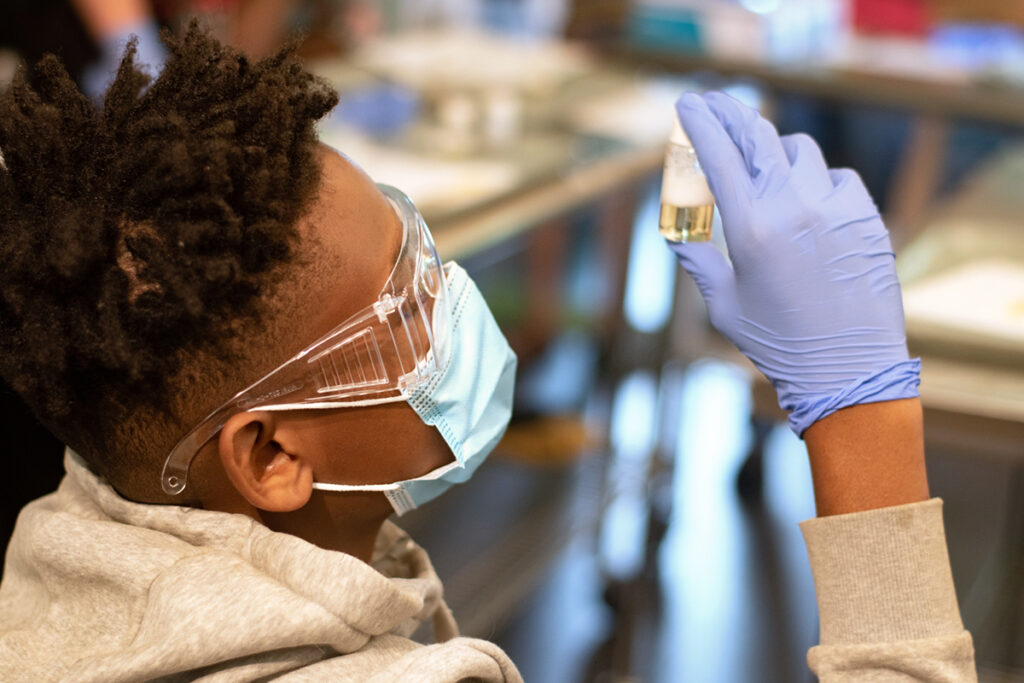 A brown-skinned person wearing lab goggles, a surgical mask, and blue vinyl gloves examines a small vial of liquid. 