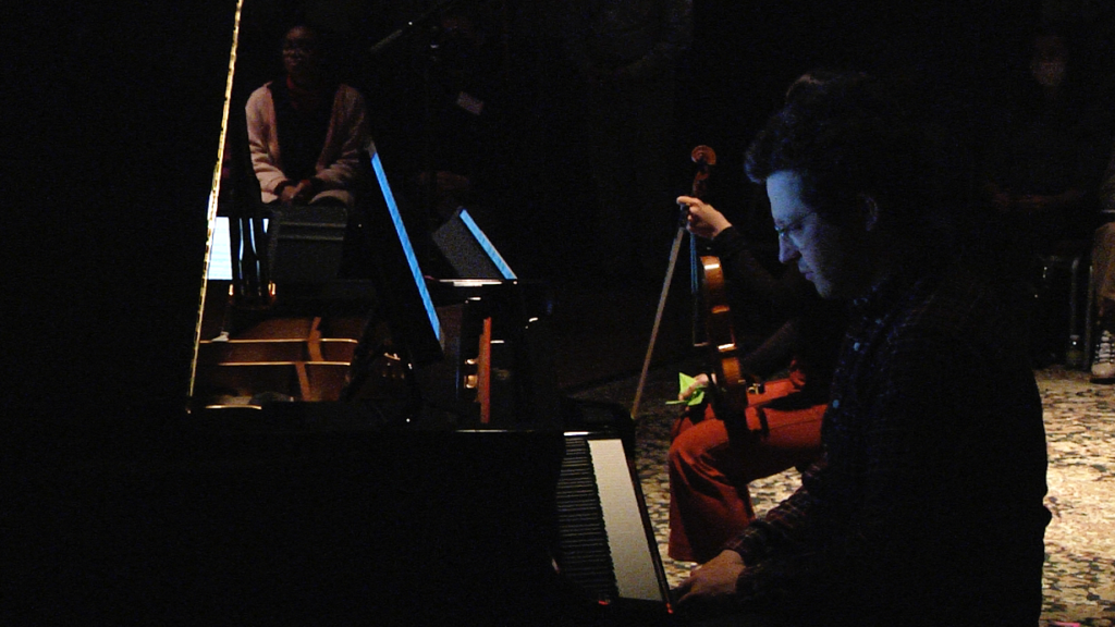 A wider shot of a person playing the piano, with audience members and fellow musicians in the background