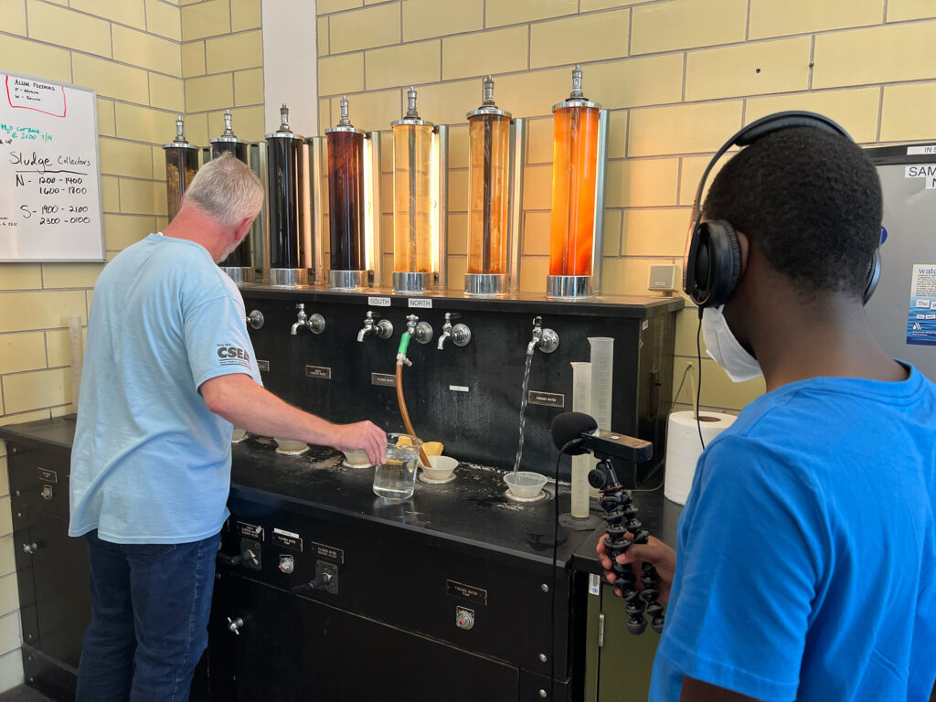William stands in front of a series of seven glass vials to demonstrate different stages of the water purification process as a water justice fellow looks on.