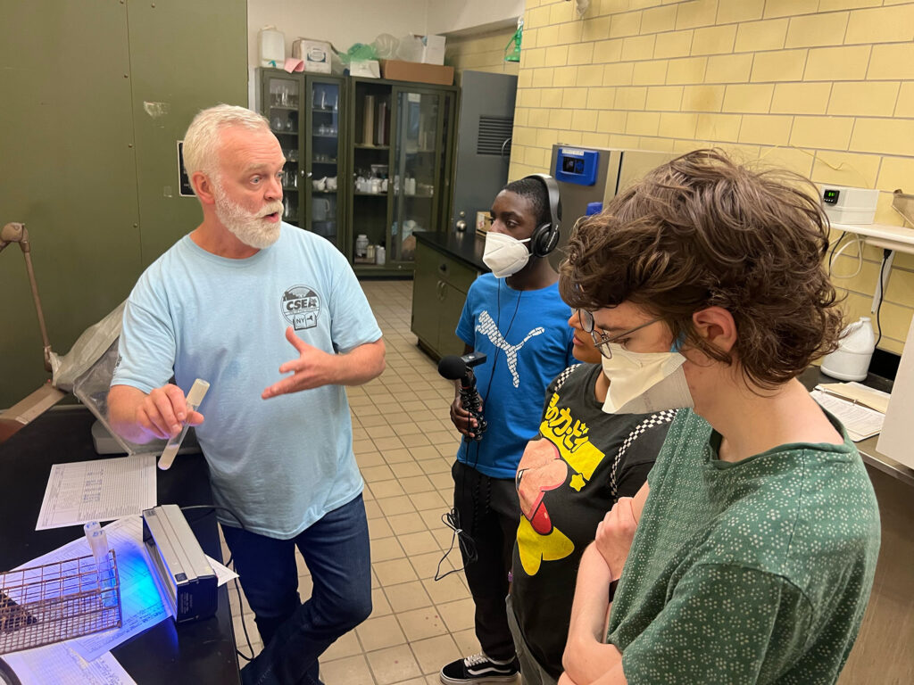 William Spillane, holding a water sample tube, speaks to three youths in the Troy drinking water plant. One of the youths records William with a microphone. 