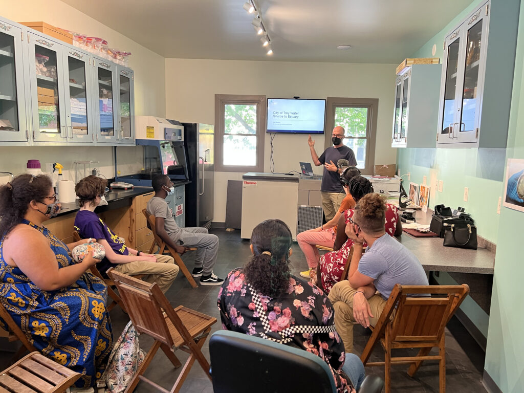 A group of attendees of various ages and ethnicities sit in the NATURE Lab space and listen as Dan gives a presentation, gesturing at a screen. 