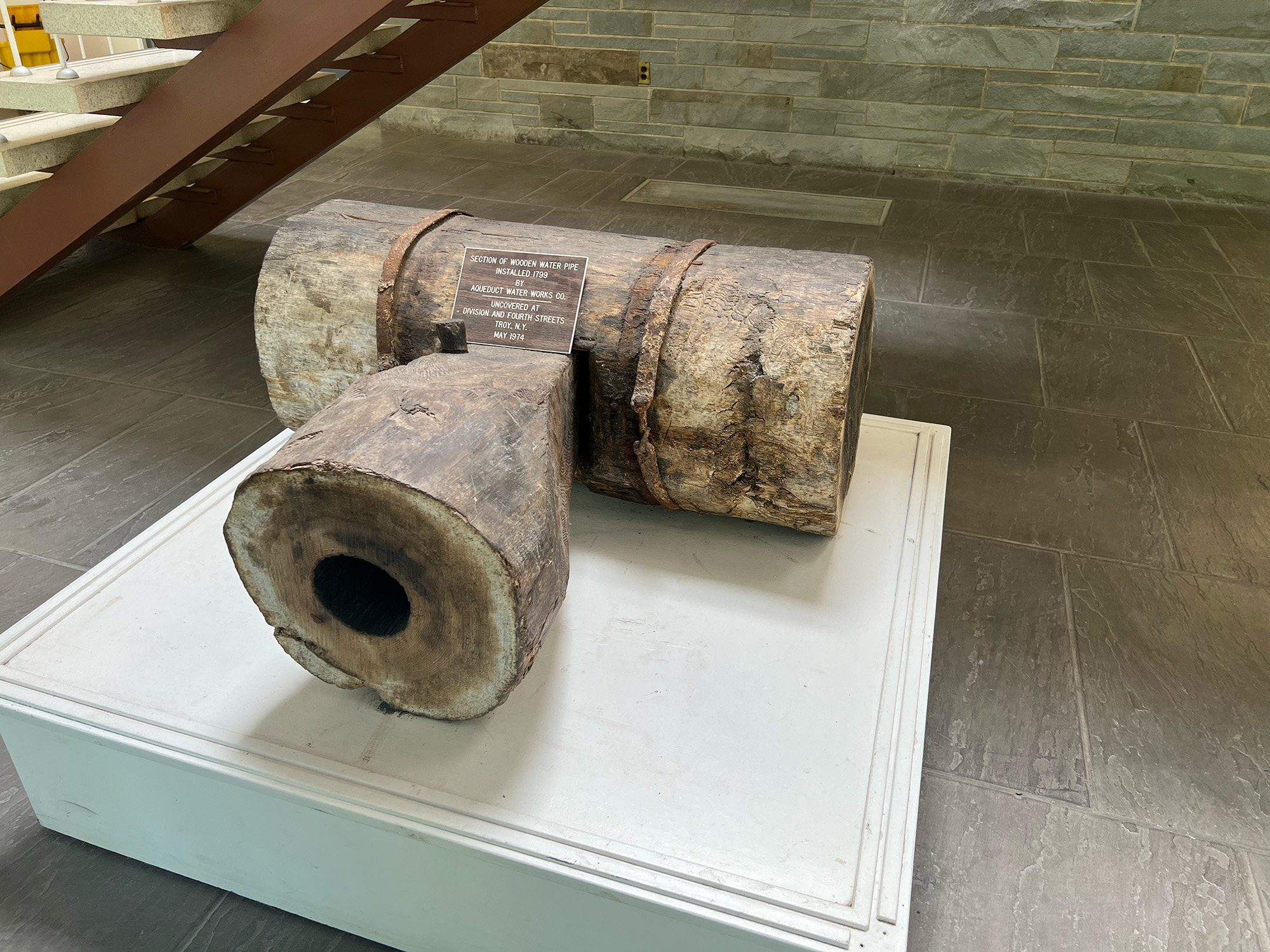 A very old water pipe joint is mounted on a square platform in the lobby of the water plant. There is a small plaque on top of the pipe.