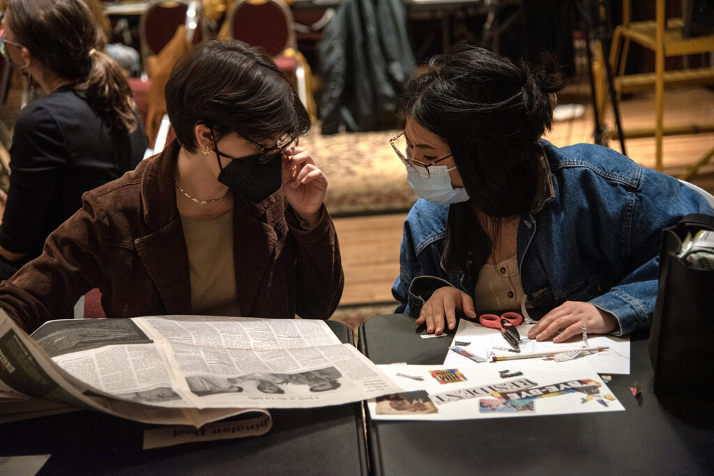 Two people sit reading a collection of newspapers and clippings, seemingly making a collage.