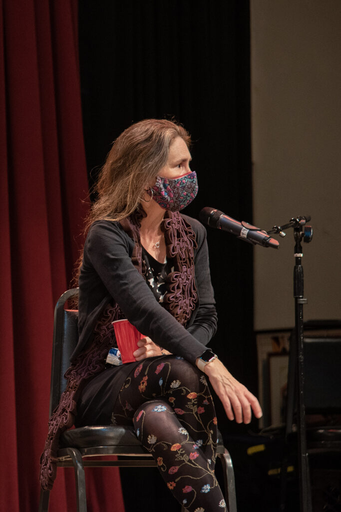 Amy Sara Carroll sitting and talking to audience. She is wearing floral pants as well as a floral mask, along with a black long sleeve.