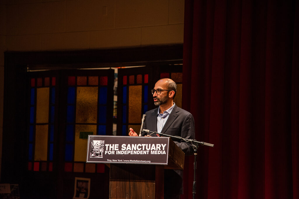 Shahan Mufti at the podium at the Sanctuary for Independent Media, talking to the crowd and wearing a blue button up with a black blazer and glasses.