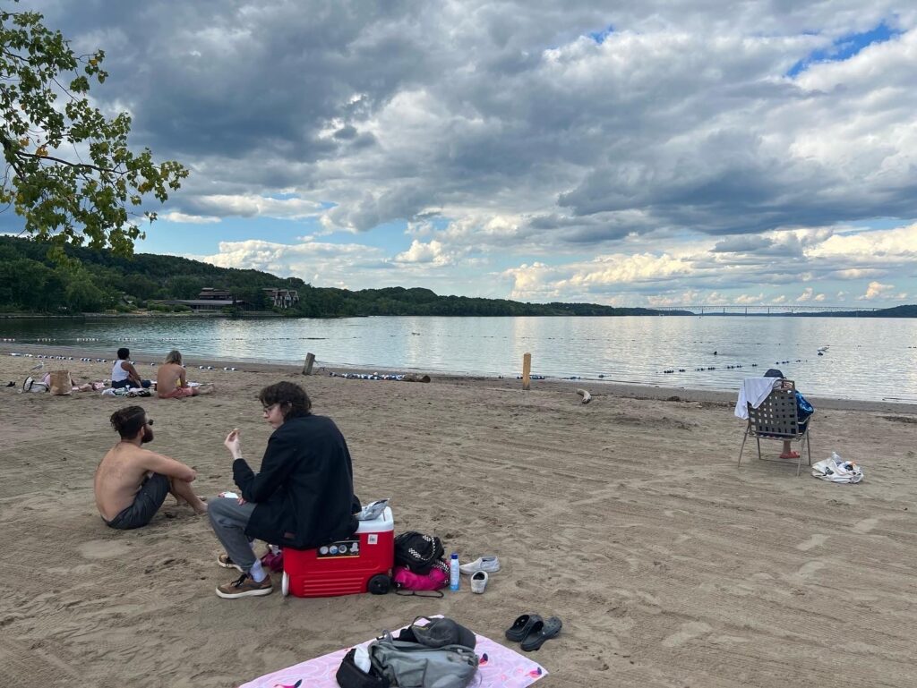Five people relax on the shore of the Kingston Point Beach on the Hudson River. The sky is mostly overcast. 