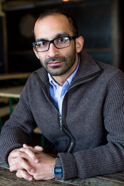 A medium-framed headshot of Shahan Mufti, a medium-skinned man wearing glasses and a grey sweater on top of a blue button up, looking into the camera with his hands intertwined resting on the table he is sitting at.