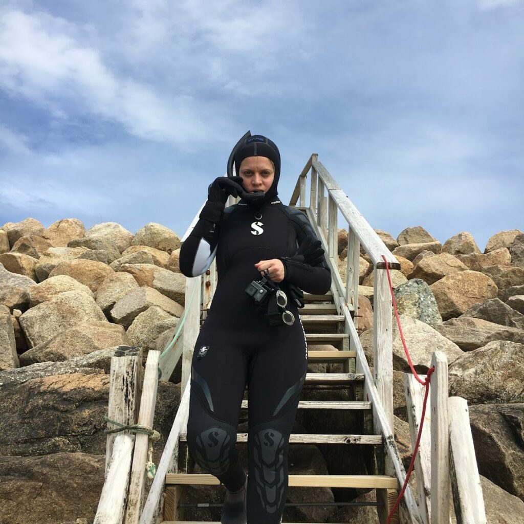 An image of Miriam Simun, a light-skinned woman, wearing a full-body wetsuit preparing for a dive, walking down a set of stairs surrounded by rocks. 