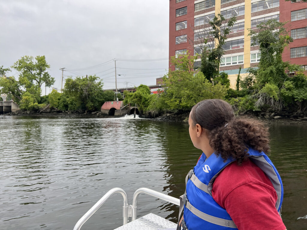 A Sanctuary youth fellow, wearing a blue life vest, looks out across the Hudson River at the opposite shoreline, which has a wastewater outflow tunnel and various trees. 