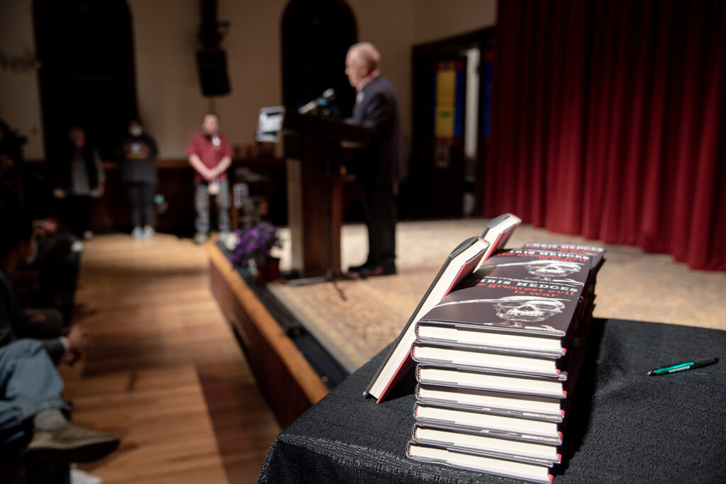 A farther shot of Chris Hedges, showcasing is latest book in the bottom right corner, stacked in several piles.
