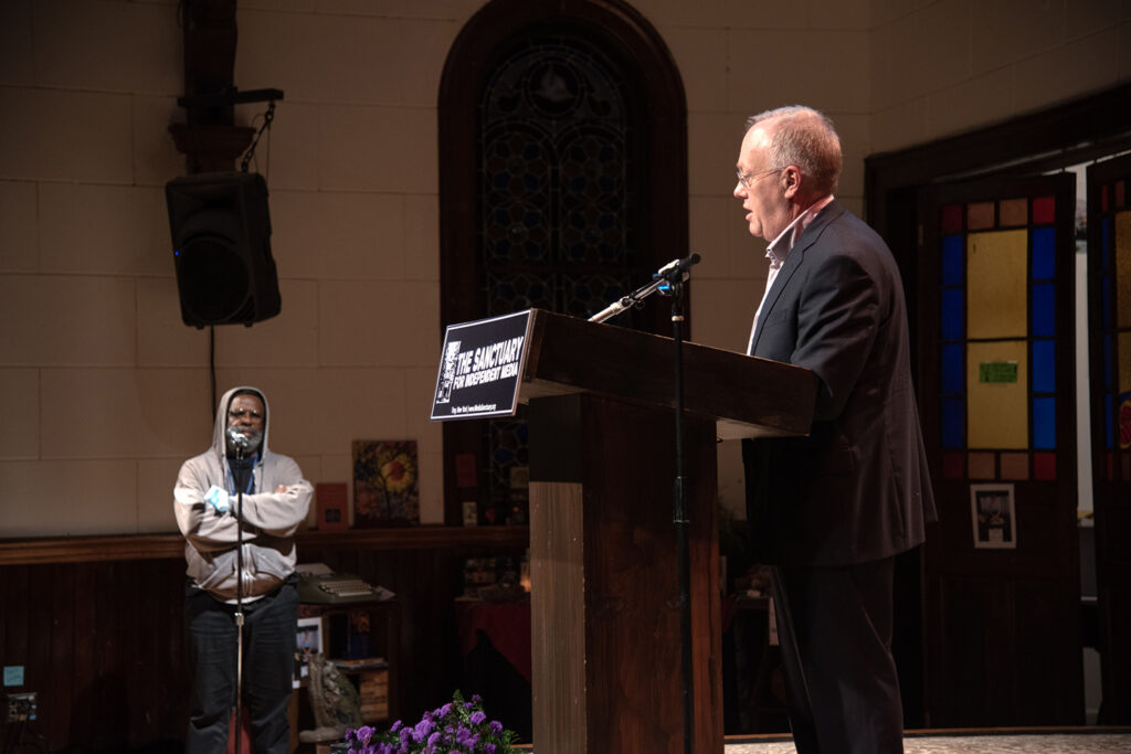 An image of Chris Hedges standing at the podium for the Sanctuary for Independent Media talking to an audience member.