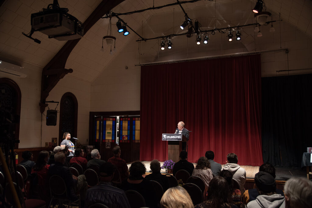 An image of Chris Hedges standing at the podium for the Sanctuary for Independent Media (farther into the audience)