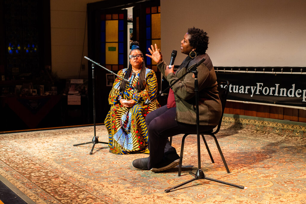 An image of Yoruba Richen and Dr. Veneilya Harden talking in front of an audience. Richen is waering a red shirt, green jacket and black pants and has short, black hair and Harden is wearing a colorful dress with her hair half up.