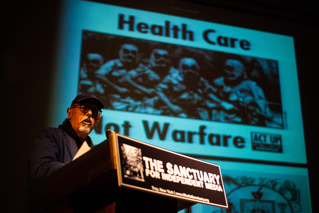 Ricardo Dominguez stands at the podium with the words Health Care Not Warfare on the slide behind him.