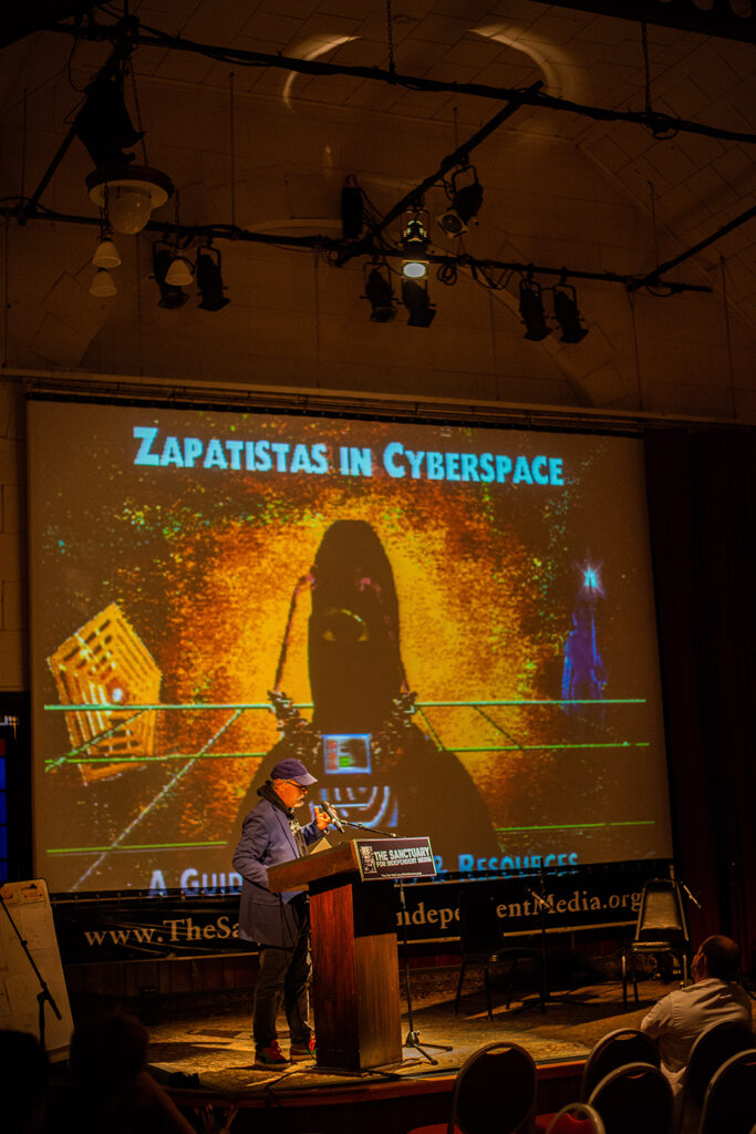 An image of Ricardo Dominquez standing before the podium for the Sanctuary for Independent Media, talking to the audience with a presentation behind him.