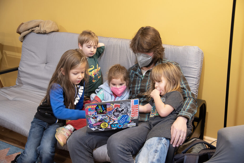 A group of children are gathered around a young adult holding a laptop, all of the children looking at whatever is on the screen. The back of the laptop is covered in stickers.
