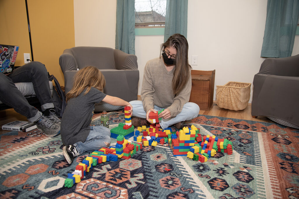 The floor is covered in colorful play bricks atop a rug, and a light-skinned person with long brown hair is playing with a younger child, also light-skinned with medium-length brown hair. They are building something on the ground. 