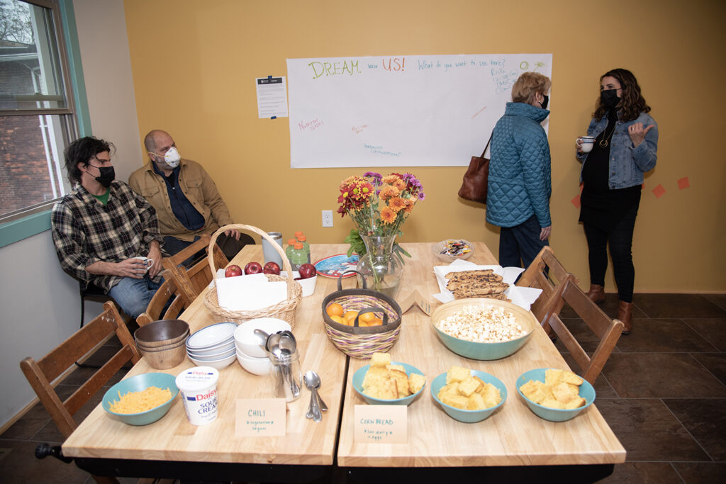 We see the table, with all kinds of snacks and food displayed, and a larger, clear image of the poster board people have been writing on. There are two people sitting int he corner, and two people standing to the right talking.