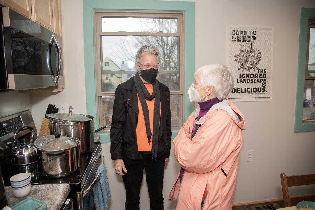 Two light-skinned people stand in a kitchen, talking in front of a stove full of pots and a window behind them. Both wearing masks.
