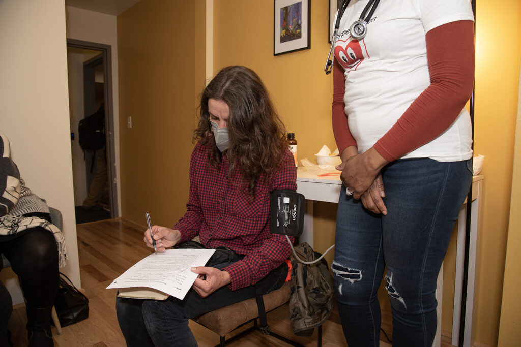 A light-skinned woman with medium-length brown hair, wearing a mask and pink and black plaid shirt, is getting her blood pressure checked and filling out a form. Standing to her right is one of the people of color from the photo on the left.