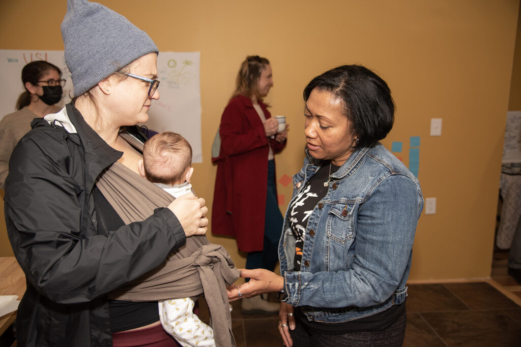 A light-skinned person with a baby in their arms is talking to a person of color, who is reaching out to touch the baby. 