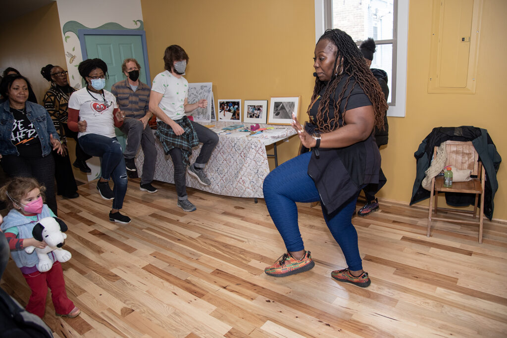 A dark-skinned woman with waist-length hair leads a group of people in a dance at the People's Health Sanctuary. 