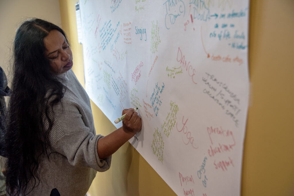 A medium-skinned person with long black hair wearing a grey sweater writing on a poster, which includes other writing of what people want to see at the People's Health Sanctuary (some words are dance, yoga, singing, etc.)