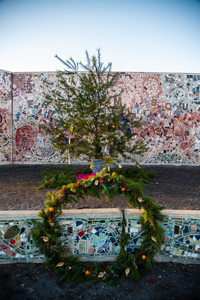 A wreathe covered in ornaments with a tree behind it, also highly decorated and both in front of a mosaic wall.
