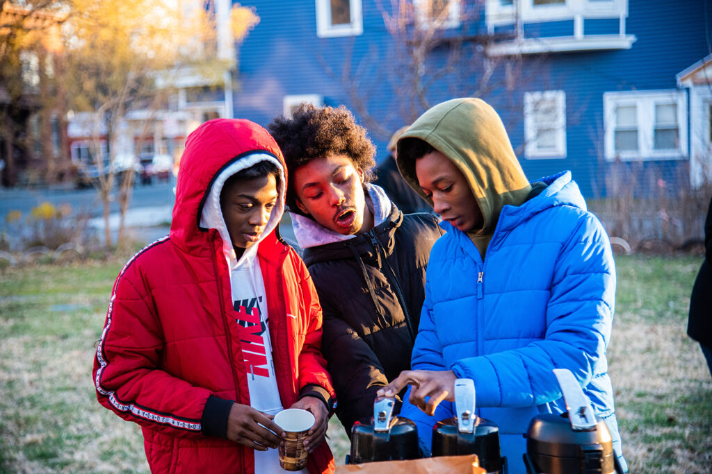 Three people or color, ranging from children to teenagers, pouring hot cocoa from a container. From left to right, they are wearing a red jacket, black jacket, and blue jacket. They stand outside a house in a front yard.