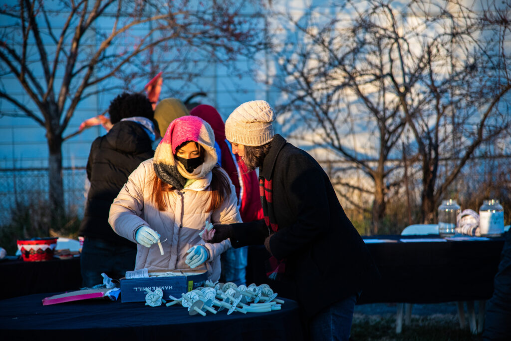 Two woman putting together the candles pictured previously, each bundled up for the cold wearing hats and jackets as they stand at a table preparing for the event. 