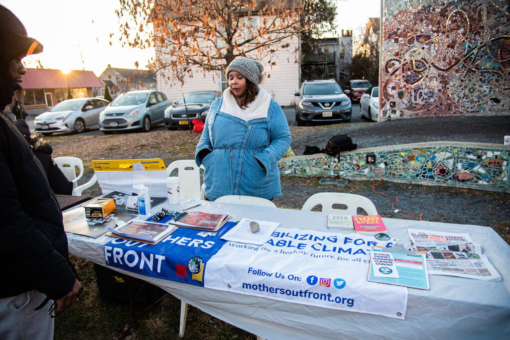 Another table held by a medium-skinned person wearing a blue jacket and grey hat, with an advertisement for Mothers Out Front and various posters/leaflets describing the organization. They are in front of a parking lot and a mosaic wall.