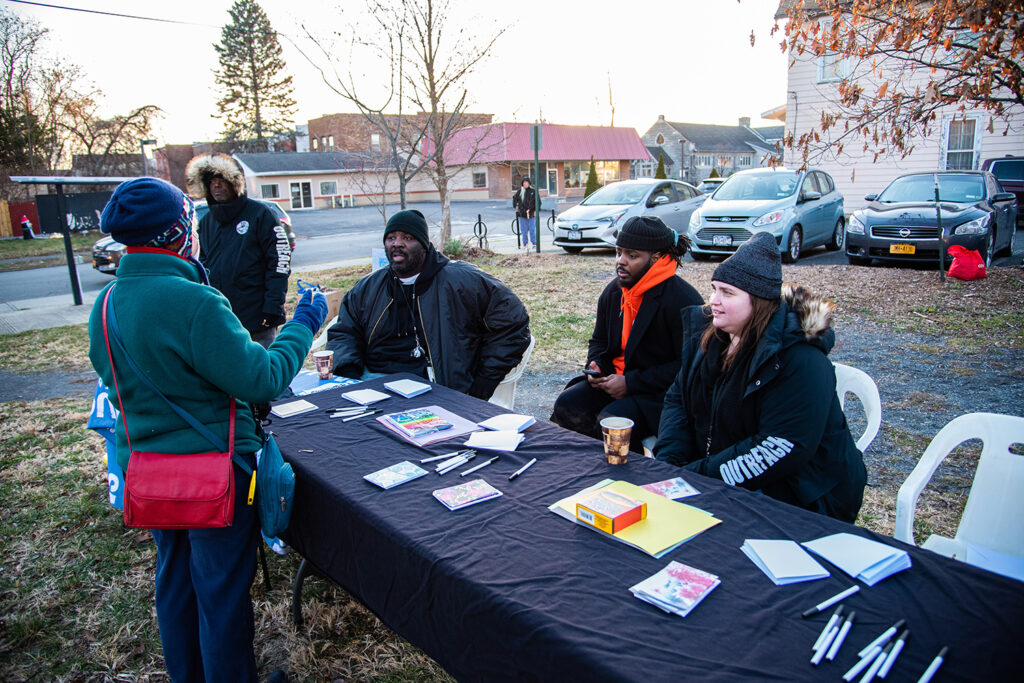 A group of people sit at a table with pens and notepads strewn across it, with three people of color and one light-skinned woman working behind and another person standing in front talking to them. There is a parking lot behind them as well as several houses and buildings.
