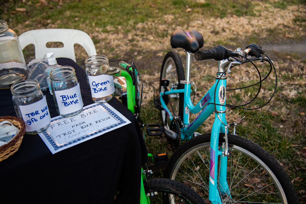 An image of two bikes, blue and green, and a table that has three jars, labeled TBR (troy bike rescue) bike gift card, blue bike, and green bike. Below these jars is a poster that reads Free Bike from Troy Bike Rescue.