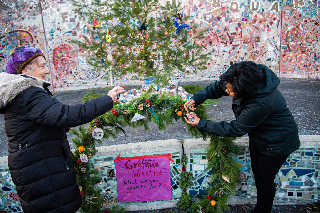 Two people (on left, light-skinned person with purple hat and black jacket, and on right, a person of color with black hair and a black jacket) putting ornaments on a wreathe. In the center is a poster that reads 2022 Gratitude Wreathe What are you grateful for?. There is also a tree behind them and a mosaic wall. 