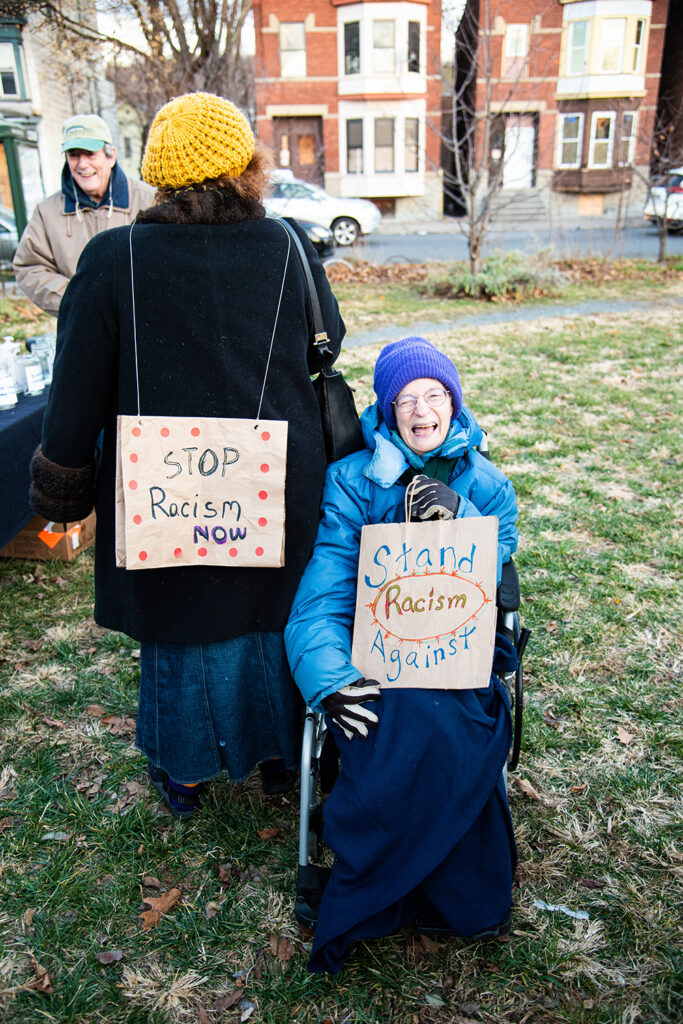 An image of several people, holding sings/ wearing signs. On the right is a light-skinned person in a wheelchair bundled up in all blue, holding a sign that reads Stand Against Racism. On their left, another person stands with their back to the camera with a sign strung across their back reading Stop Racism NOW, wearing a black jacket and yellow hat. There are also people in the background, as well as houses and more tables for the event. 