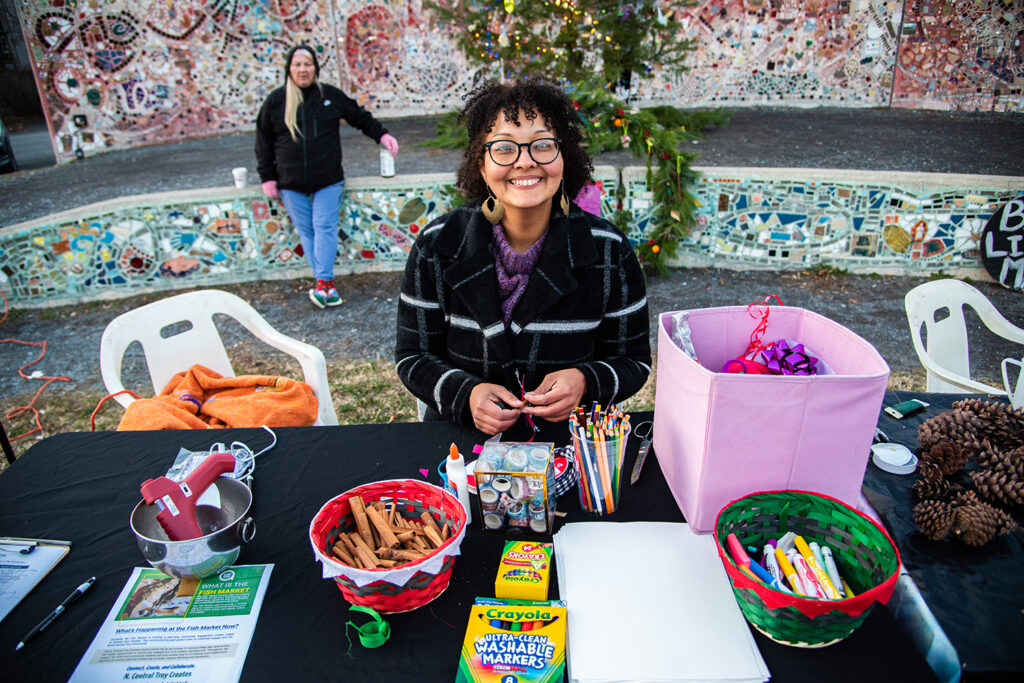 An image of a smiling, light-skinned person with short curly hair standing behind a craft table, which is filled with crayons, markers, and other supplies. Behind them stands another person and a mosaic scene with more decorated trees.