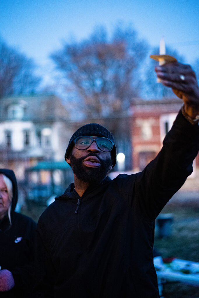 A single-shot of a person of color holding up a candle in their left hand for the camera, wearing glasses and a beanie with a black jacket.