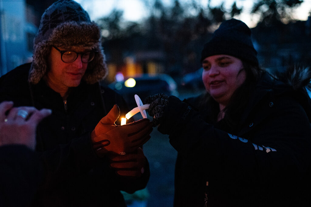 The person on the right is using their candle to light the person on the left's. Both are wearing hats and jackets and it is becoming dark outside.